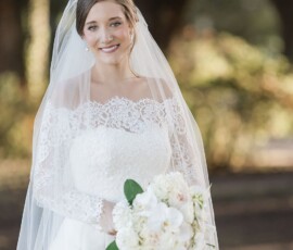 Bride in white lace dress and veil holding a bouquet outdoors, smiling at a joyful Louisiana wedding with trees behind her.