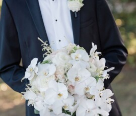 Man in black tuxedo with white boutonniere holds white orchid and rose bouquet outdoors at Louisiana wedding.