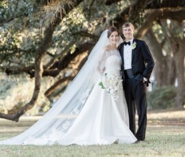 Smiling bride in white gown and groom in tuxedo stand under sunlit trees before their wedding ceremony.