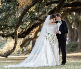 Bride in white gown and groom in tux share an intimate moment under large trees at their Louisiana wedding.