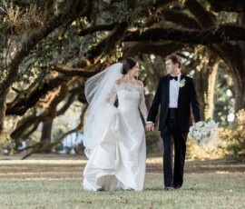 Bride in white gown and groom in black tuxedo walk hand-in-hand beneath sunlit trees, classic Gulf South wedding.