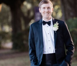 Smiling man in black tuxedo with boutonnière stands outdoors, trees and sunlight evoking a classic Louisiana wedding.