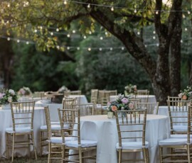 Elegant outdoor wedding reception with white-clothed tables, gold chairs, floral centerpieces, and string lights.