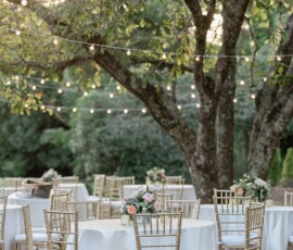 Elegant outdoor wedding with round white-clothed tables, gold chairs, floral centerpieces, and string lights among leafy trees.