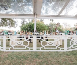 Outdoor wedding bar with bartender, bottles, floral decor, and guests mingling under a clear tent.