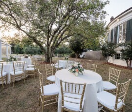 Elegant outdoor wedding setup with round tables, gold chairs, and flower centerpieces beside a white house with green shutters.