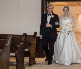 Bride in white gown with flowers walks down church aisle, escorted by man in tuxedo; another woman holds her veil.