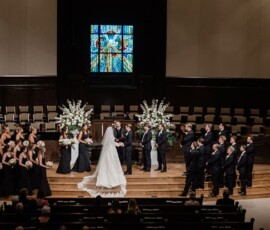 Bride and groom with wedding party at church altar, floral arrangements and stained glass window in background, guests watching.