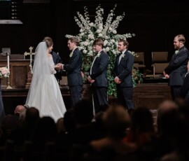 Bride and groom exchange vows at the altar, surrounded by their wedding party and white floral arrangements in Louisiana.