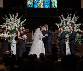 Bride and groom kiss at the altar, surrounded by bridesmaids in black, groomsmen in suits, and white floral arrangements.