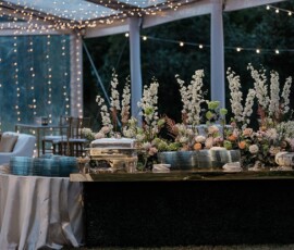 Buffet table with flowers and plates under string lights at an outdoor New Orleans wedding beneath a clear tent.