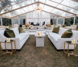 Stylish outdoor lounge under a clear tent with white sofas, fire pit table, gold accents, and checkered dance floor for weddings.