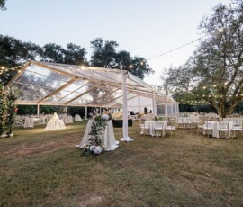 Transparent tent with string lights over tables and chairs for a Louisiana wedding, surrounded by trees in early evening.
