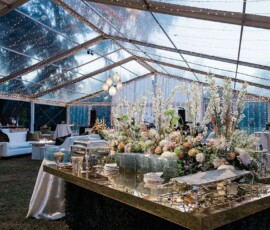 Elegant buffet table with flowers under a clear tent, string lights, and guests at a stylish Gulf South wedding reception.