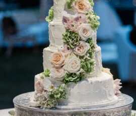 Elegant four-tiered white wedding cake with pastel roses and green hydrangeas on a silver stand, softly lit.