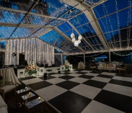 Clear tent with string lights over a black and white checkered dance floor, floral decor, tables, and glowing orbs for a wedding.