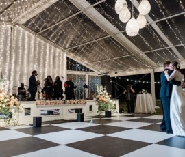 Bride and groom share their first dance on a checkered floor under a tent with lights and flowers as the band sets up.
