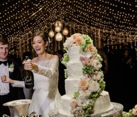 Smiling bride pops champagne by her groom and wedding cake under string lights at a joyful Louisiana wedding reception.