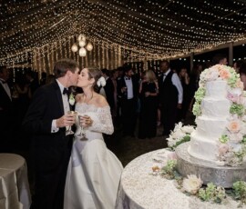 Bride and groom kiss with drinks at their Louisiana wedding, string lights overhead, white floral cake nearby, guests celebrating.