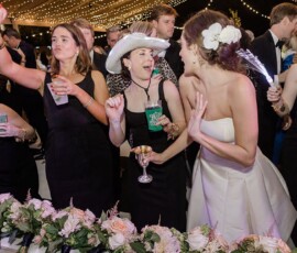 Bride in white laughing with bridesmaids in black at lively Louisiana wedding, drinks and string lights overhead.