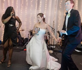 Bride and groom dance on stage holding hands, with a singer, string lights, and live band at a Louisiana wedding.