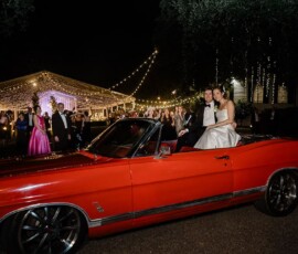 Bride and groom in a red convertible at night, smiling at wedding guests under string lights outside a lit-up New Orleans tent.