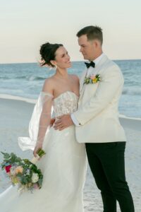 Bride and groom in white smile on a Louisiana beach, waves behind them. Bride holds bouquet; groom wears white jacket and bow tie.