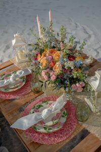 Elegant outdoor wedding table on sand with floral plates, green glasses, pink decor, and a colorful bouquet.