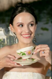 A smiling woman in a white strapless dress holds a floral teacup outdoors, sunlight on her face—ideal Gulf South wedding inspiration.
