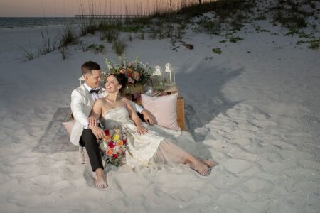 Barefoot newlyweds sit on a decorated beach blanket, smiling at sunset over the Gulf South ocean.