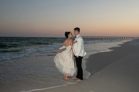 Bride and groom barefoot on Louisiana beach at sunset, facing each other with waves and a pier in the background.