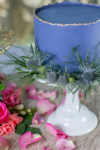 Blue cake with gold edge on white stand, topped with thistle and greenery. Pink roses and petals decorate the table.