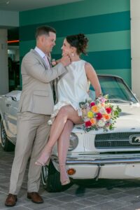 Bride and groom pose by a classic white car in New Orleans, with bride on hood holding bouquet and groom touching her chin.