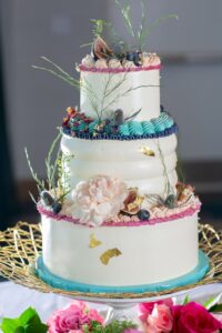 Elegant three-tiered white cake with pink, blue, and gold accents, figs, berries, florals, on a gold stand with pink roses.