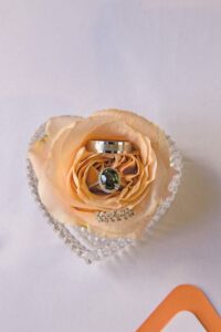 Peach rose in heart-shaped glass dish holds wedding band and gemstone ring on petals, with rhinestone accent.