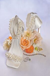 Pearl-adorned white heels with peach and white flowers, jeweled bracelet, ring, and gold earrings on a light background.