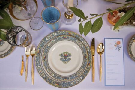 Elegant Louisiana wedding table with blue and gold plates, gold cutlery, glasses, menu, and greenery on a white cloth.