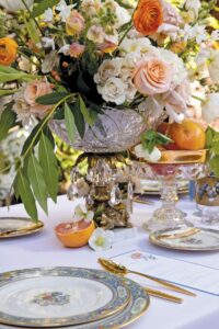 Elegant outdoor wedding table with flowers, crystal bowls of fruit, gold flatware, and vintage floral plates on white linen.