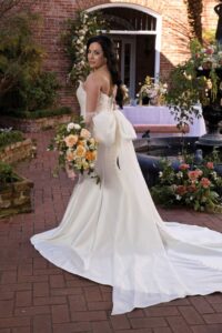 Bride in strapless white gown with bow holds peach and white bouquet on brick path, surrounded by lush floral greenery.