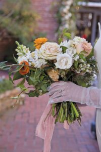 Person in sheer gloves holding bouquet of white, peach, and orange flowers with greenery; blurred brick path in background.