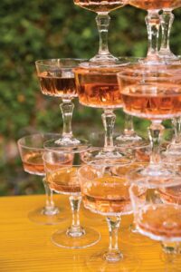 Champagne tower with rosé-filled glasses on a yellow table, greenery blurred in background—romantic Gulf South wedding inspiration.