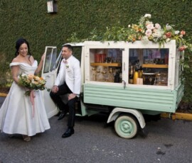 Bride and groom smile by a mint-green drink cart with flowers and bottles, set against an ivy-covered wall—wedding inspiration.