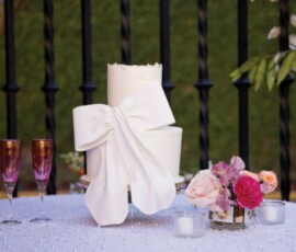 Elegant white two-tiered cake with fondant bow, flanked by pink champagne flutes, flowers, and candles—Gulf South wedding décor.