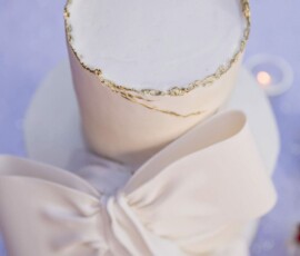 Elegant white two-tier cake with gold edging and fondant bow on lilac tablecloth, perfect for a New Orleans wedding.