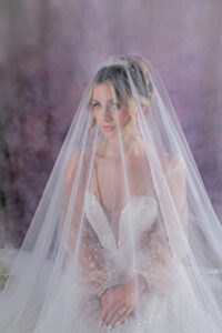 Bride in sparkling white gown and veil sits with hands folded, gazing sideways against a softly blurred pastel purple background.
