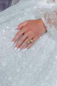 Hand with long white nails and a gold gemstone ring rests on a sheer, lace sequin dress.