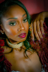 Woman with glowing skin, bold eye makeup, and dark red lipstick poses in pearls and red attire against a deep red background.