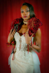 Confident woman in a lace gown with red floral shoulder piece, gold rings, and sparkling choker poses against red background.