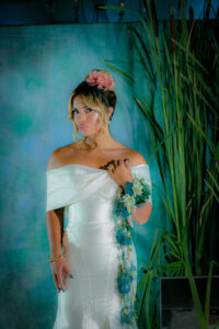 Woman in off-shoulder white gown with floral accents stands before green backdrop and tall grass, facing the camera.