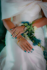 Bride in a white dress with blue floral nails, gold jewelry, and a white and green bouquet on her lap.